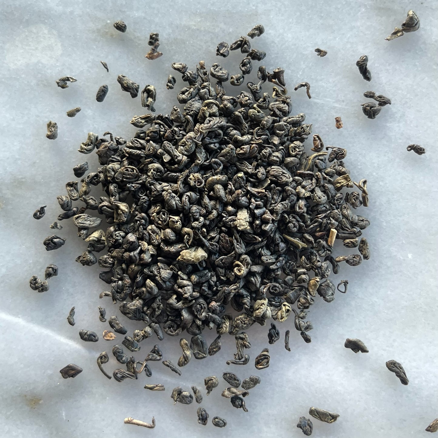En liten tehög med det gröna teet Gunpowder, liggandes på en vit marmorbricka där man kan se teets färg och struktur
