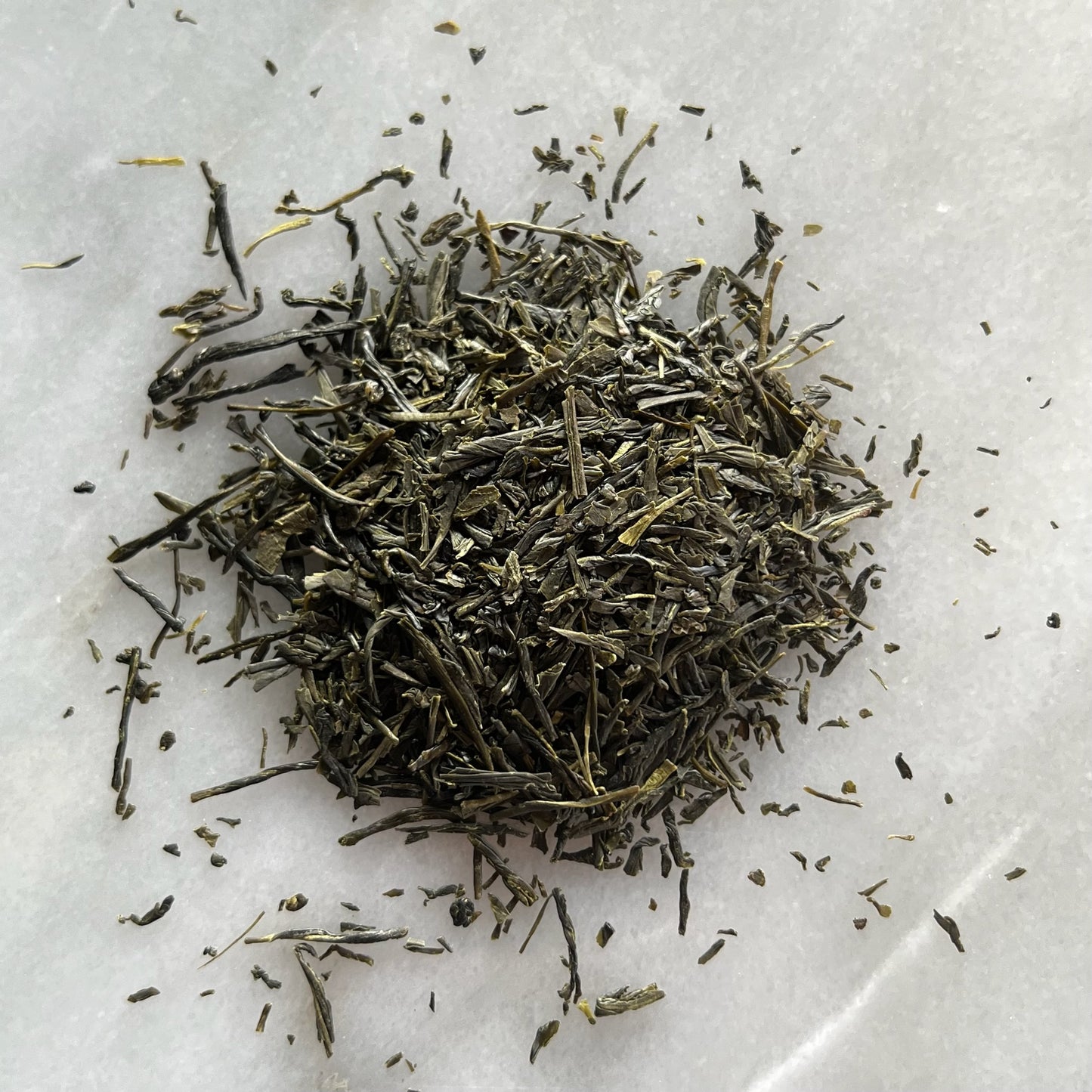 En liten tehög med det gröna teet Japansk Sencha, liggandes på en vit marmorbricka där man kan se teets färg och struktur