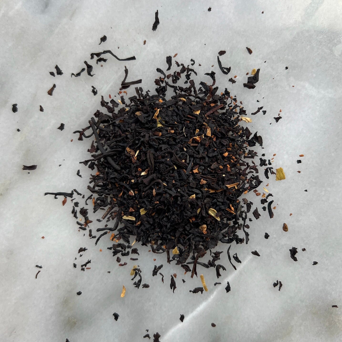 En liten tehög med det svarta teet Mys Pys (Needwoods läckerhet), liggandes på en vit marmorbricka där man kan se teets färg och struktur