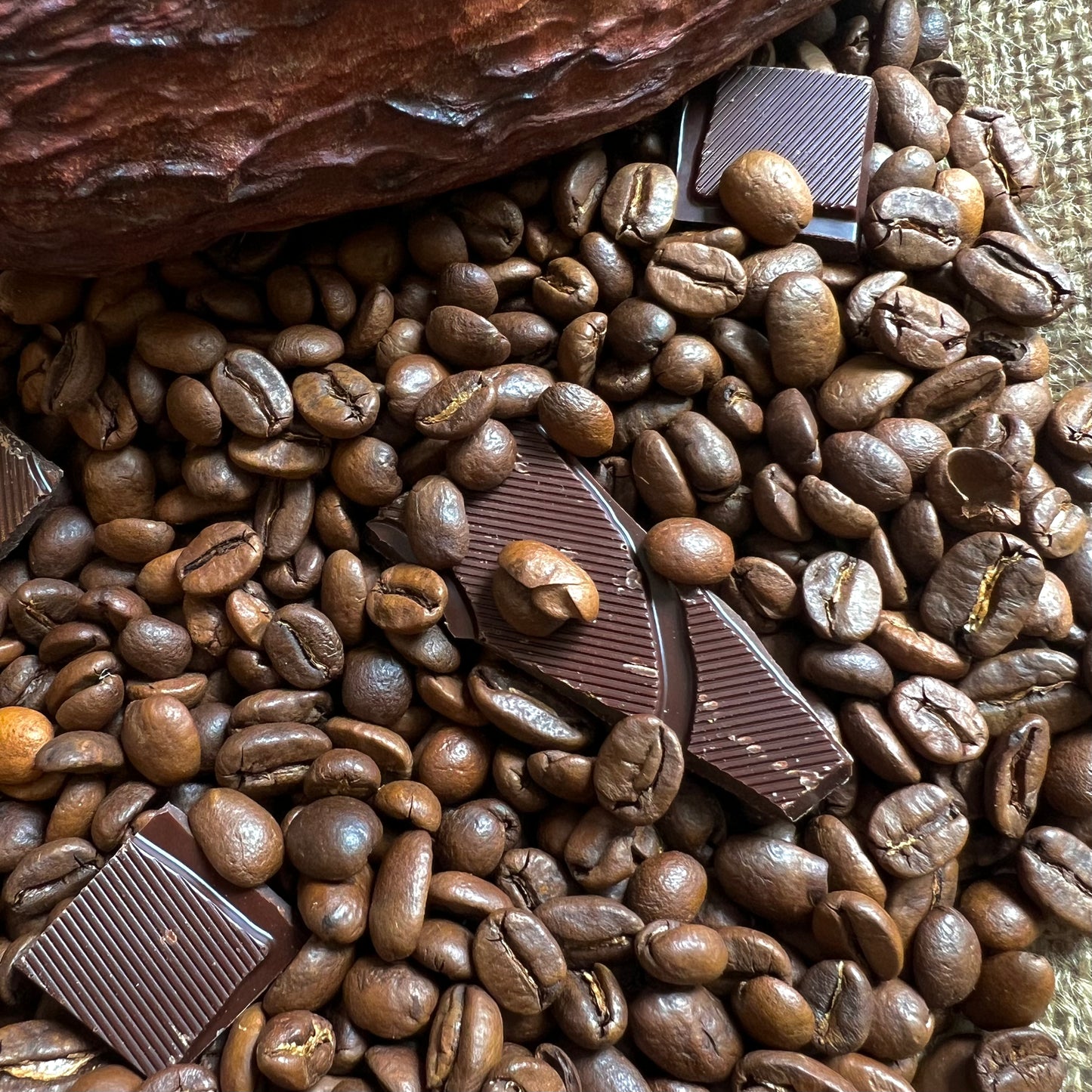Närbild av kaffebönor och chokladbitar på en jutesäck med en kakaofrukt i hörnet.