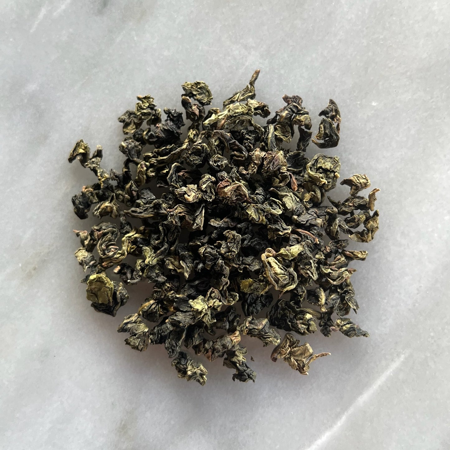 En liten tehög med det gröna teet Oolong Tie Kuan Yin Superior, liggandes på en vit marmorbricka där man kan se teets färg och struktur